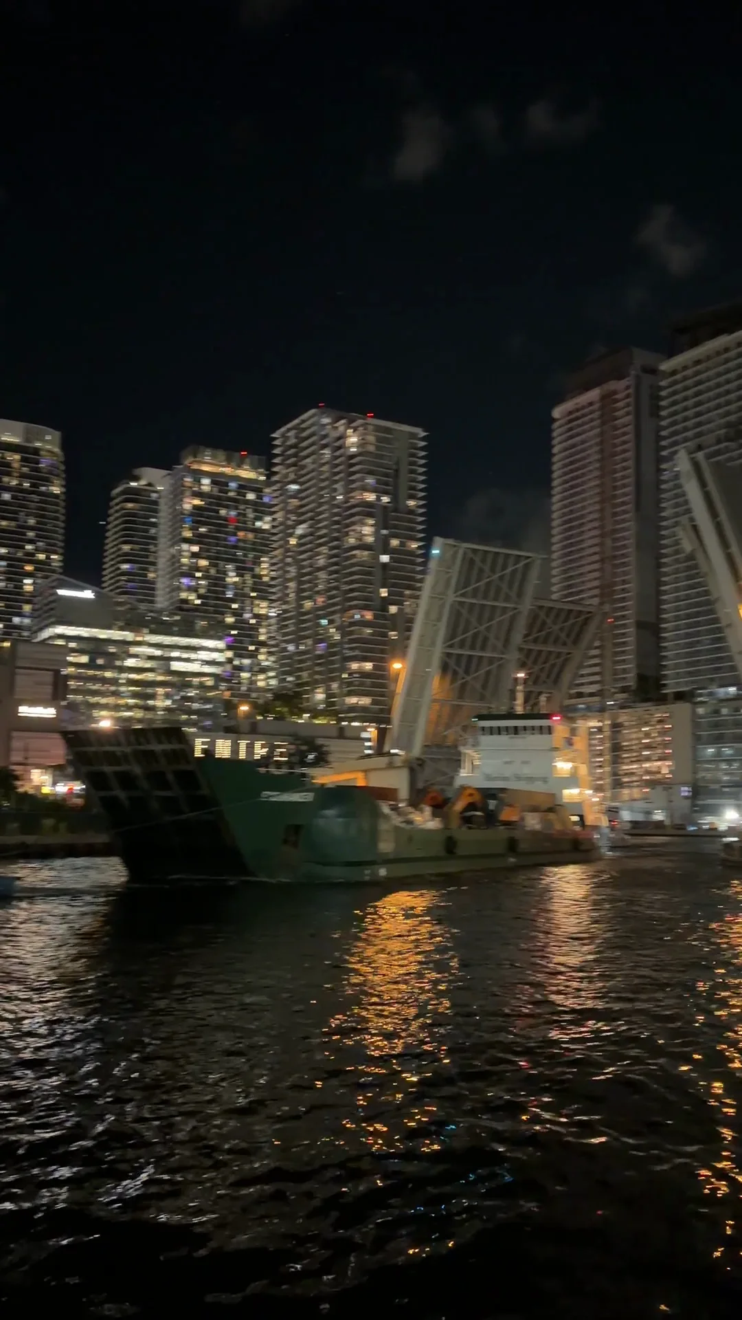 A Bimini Shipping cargo ship passing under the raised Brickell Avenue bridge at night, the Miami skyline lit behind it